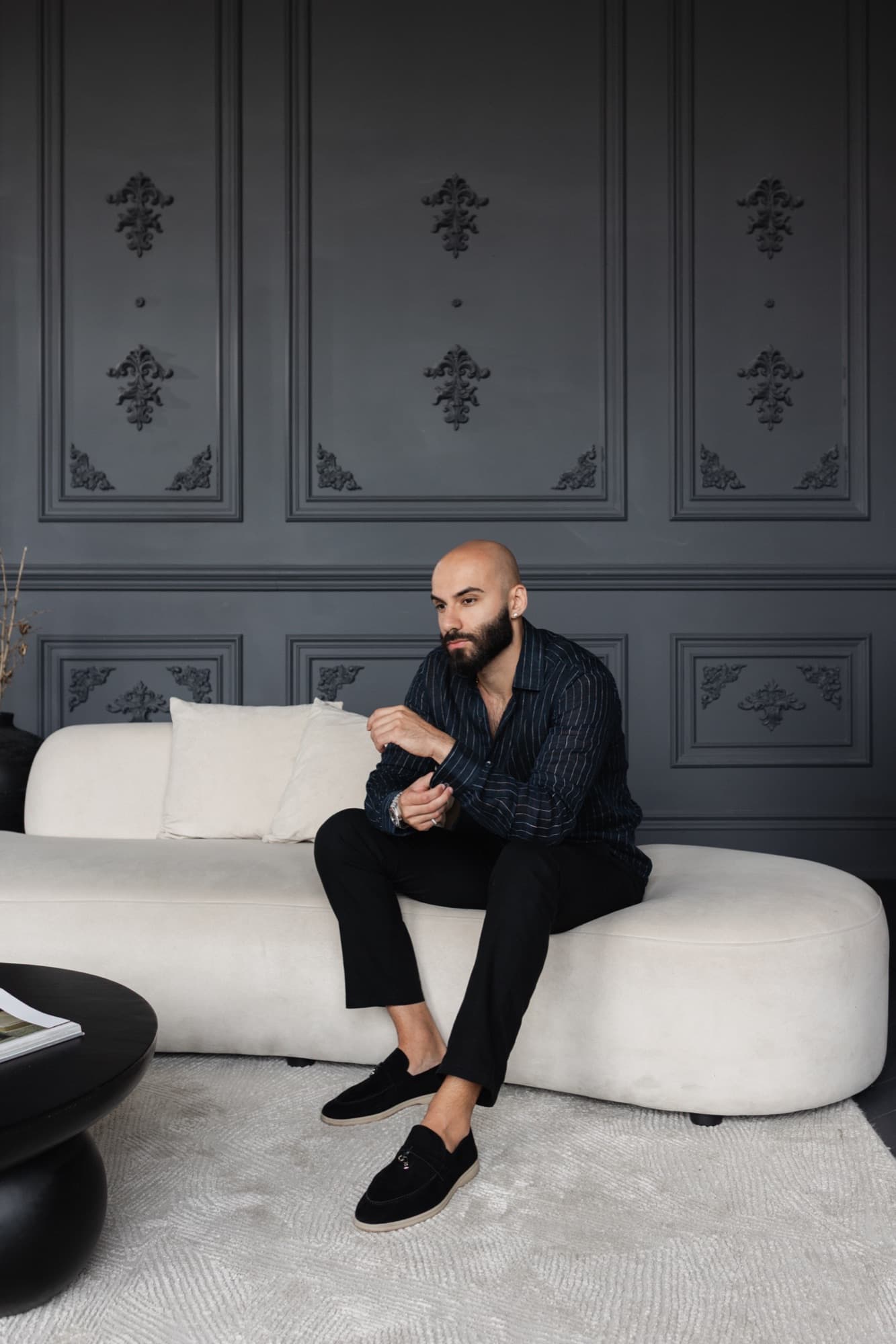 Navid Rashid seated on a cream sofa in black, against a grey panelled wall