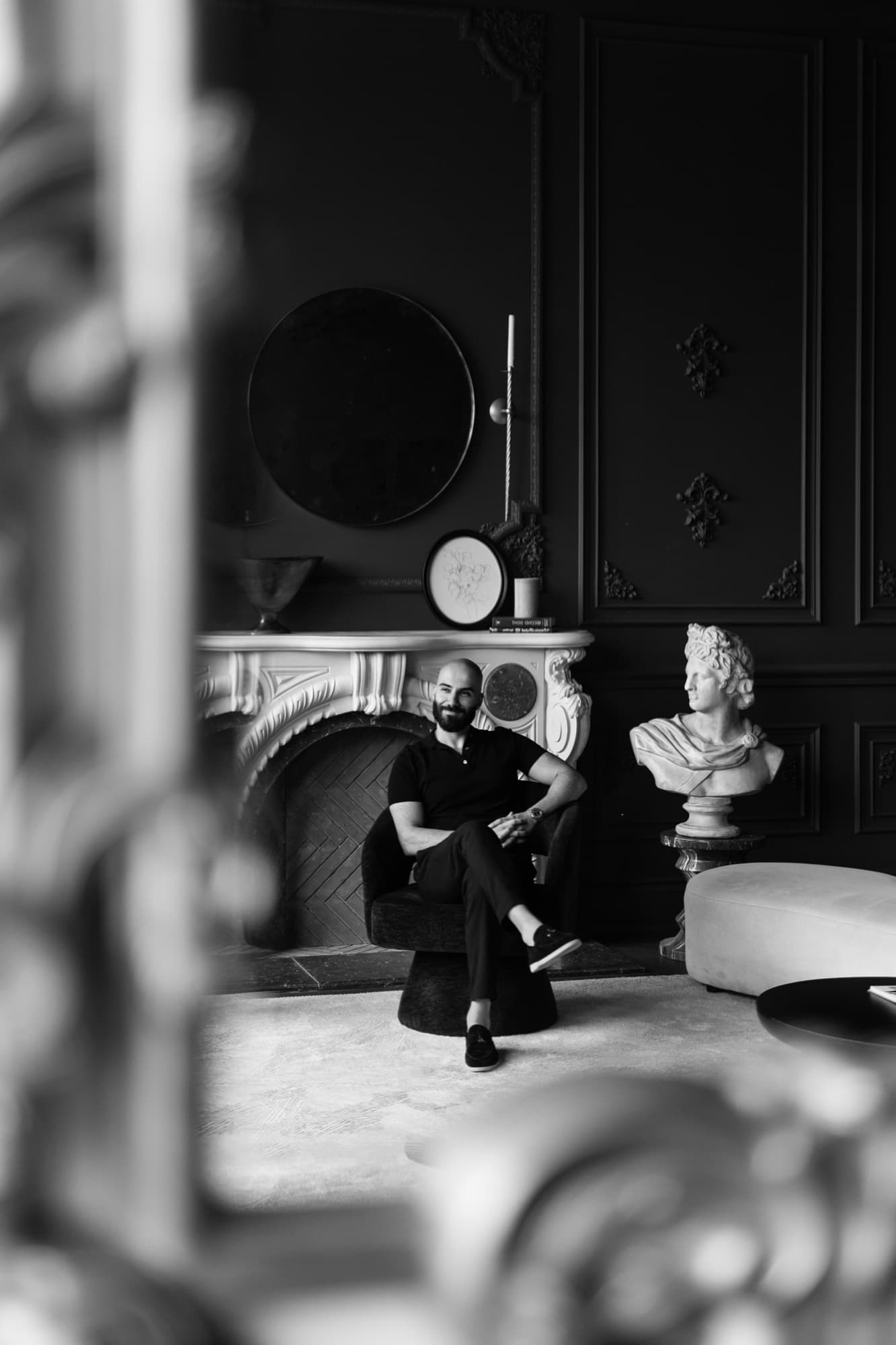Black and white portrait of Navid Rashid seated in a room with a marble bust and fireplace