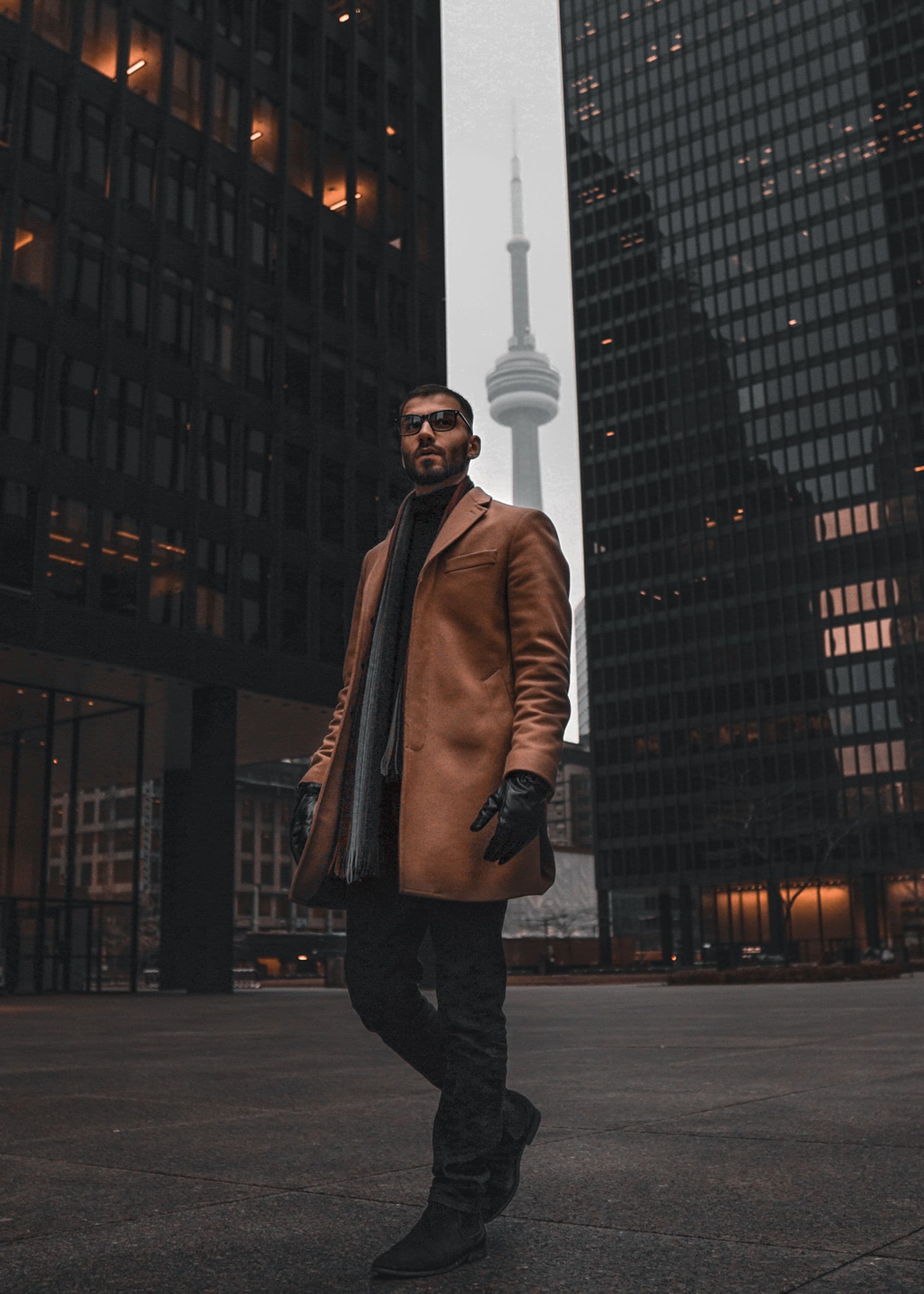 Navid Rashid in a camel coat walking in downtown Toronto with the CN Tower behind him